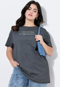 Grå kortærmet T-shirt med teksten "VELKOMMEN TIL MIN FEST" i glitrende bogstaver, parret med skinnende blå palietbukser og en blå håndtaske.