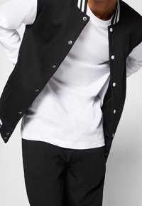 Giacca varsity nera con dettagli bianchi, bottoni a pressione e colletto a righe indossata sopra una t-shirt bianca. Design semplice e casual; materiale in misto cotone.