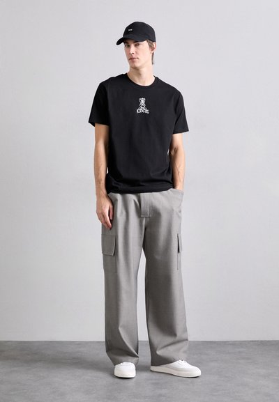 T-shirt en coton noir avec impression graphique, associé à un pantalon cargo large gris clair avec poches latérales et des baskets blanches.