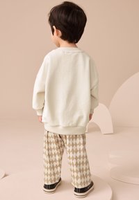 Kermanvärinen ylisuurikokoinen collegepaita resorilla varustetuilla hihoilla, yhdistetty beigeihin timanttikuvioisiin housuihin ja mustiin lenkkareihin. Lapsi kuvattu takaa päin.