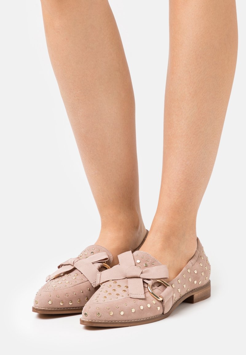 Svetle růžové semišové baleríny s mašlí, zlatými nýty a kulatou špičkou. Design zahrnuje malý podpatek a strukturovaný povrch.