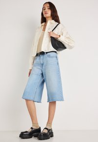 Jasna, teksturowana krótka koszula noszona na beżowym topie, zestawiona z luźnymi, jasnoniebieskimi bermudami z denimu oraz czarnymi butami na platformie.