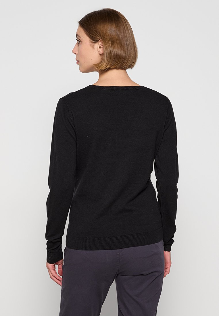 Femme aux cheveux bruns courts, portant un pull ajusté à manches longues noir et un pantalon sombre, de dos contre un fond clair uni.