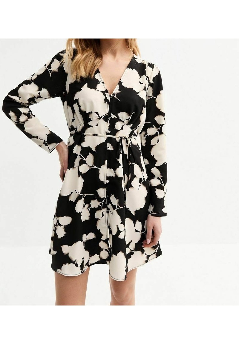Femme portant une robe portefeuille florale blanche et noire à manches longues et ceinture nouée, debout avec une main sur la hanche sur fond blanc.