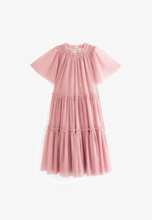 Ragazza giovane che indossa un vestito rosa a balze, trasparente, con maniche corte svolazzanti e motivo a pois glitterato, tenendo l'orlo del vestito.