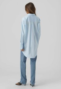 Camicia azzurro chiaro con bottoni, dal taglio lungo e rilassato, abbinata a jeans in denim chiaro e sandali neri con tacco. Tessuto morbido, texture liscia.