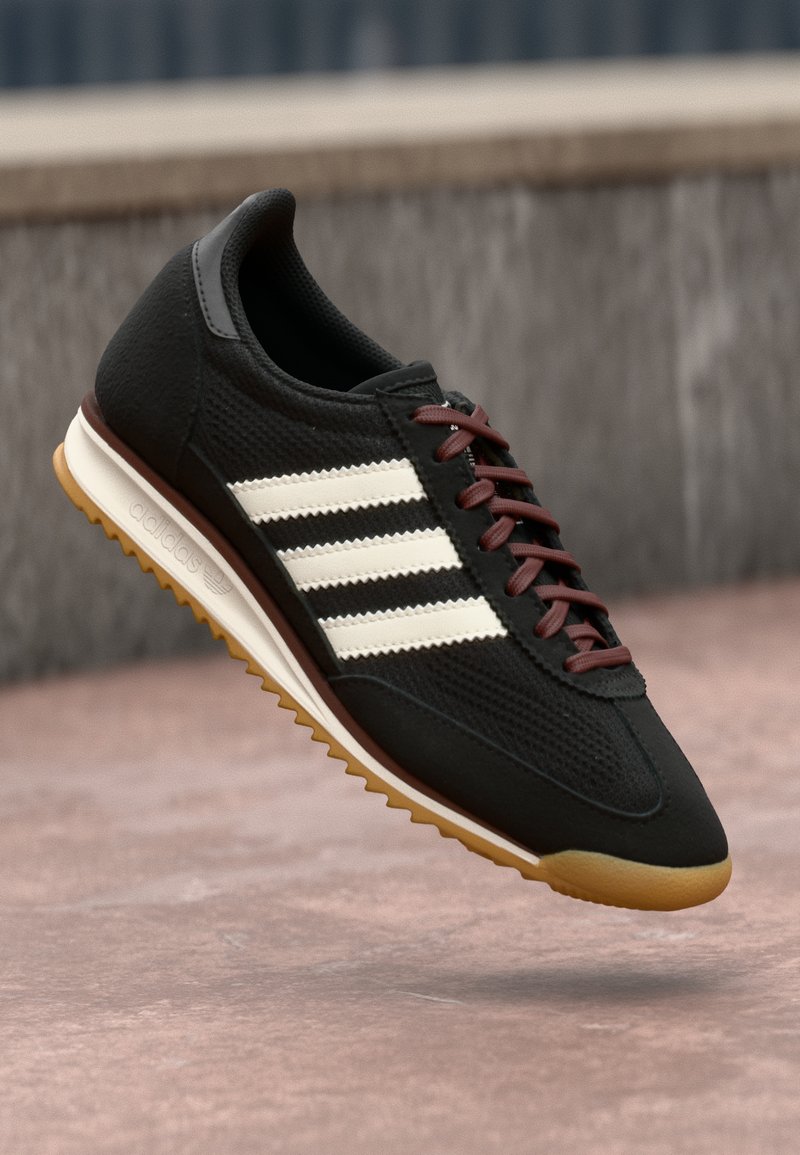 Black athletic shoe with a textured upper, three white stripes, brown laces, and a rubber gum sole. Modern design with low-profile silhouette.