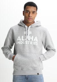 Sudadera gris con estampado blanco de "ALPHA INDUSTRIES", capucha con cordón, bolsillo canguro, puños y dobladillo acanalados. Textura de tela suave.