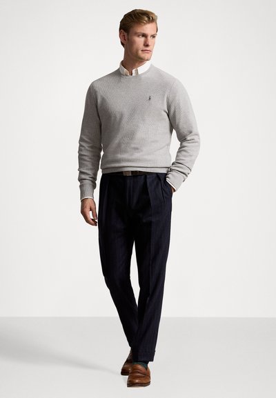 Pull en tricot gris clair avec un col ras du cou, associé à un pantalon à rayures fines noir. Des mocassins en cuir marron complètent la tenue.