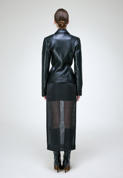 Femme aux cheveux en chignon portant une veste en cuir noire cintrée, une jupe noire transparente et des bottes noires à talons hauts, debout de dos sur un fond uni.
