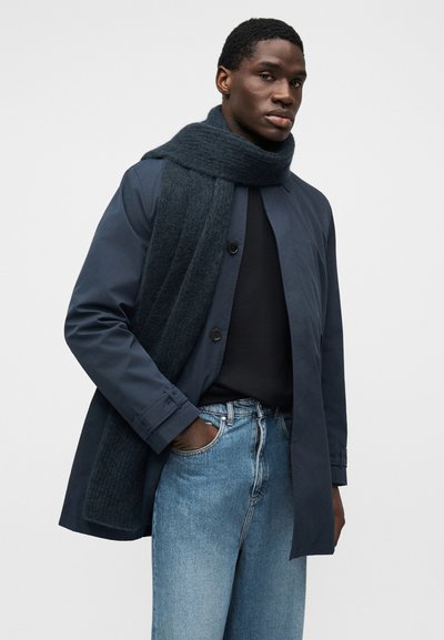 Μπλε παλτό με κουμπιά, συνδυασμένο με ένα σκούρο κασκόλ με υφή, ένα μαύρο πουκάμισο και ανοιχτό μπλε τζιν. Το ύφασμα φαίνεται λείο και δομημένο.