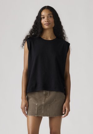 Γυναίκα με μακριά σγουρά μαλλιά, φοράει αμάνικο μαύρο τοπ και καφέ μίνι φούστα από τζιν, στέκεται μπροστά σε απλό φωτεινό φόντο.