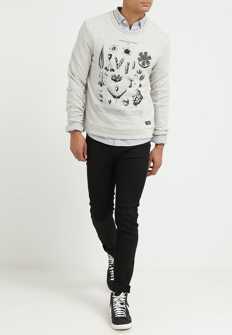 Sudadera gris con estampado botánico que presenta diversas ilustraciones negras, puños y dobladillo acanalados. Combinada con pantalones negros y zapatillas.