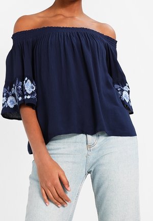 Marineblå off-shoulder bluse med elastisk halskæde, broderede blomsterdetaljer på ærmerne og afslappet pasform, kombineret med lyse denimjeans.