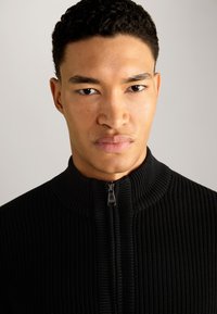 Schwarzer gerippter Reißverschluss-Pullover mit hohem Kragen und silbernem Reißverschluss. Die Textur ist gewoben und das Design hat eine figurbetonte Silhouette.