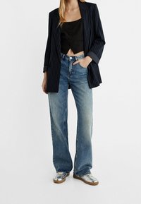 Zwart cropped topje met knoopdetails, navy pinstripe oversized blazer, vervaagde blauwe wijde jeans en glanzende zilveren sneakers met bruine zolen.
