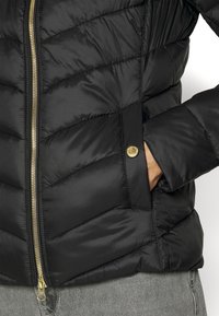 Schwarze Pufferjacke mit gesteppter Optik, goldenem Reißverschluss und Druckknopf-Taschen. Hat einen hohen Kragen und eine glatte, glänzende Textur.