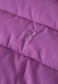 Lila gesteppter Stoff mit einem gestickten Logo, das einem kleinen "n" mit Punkt ähnelt, und hebt die Textur und Materialdetails hervor.