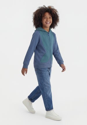 Barn med krøllete hår som smiler, går, iført blå og teal genser med glidelås, blå jeans og hvite høye joggesko på hvit bakgrunn.