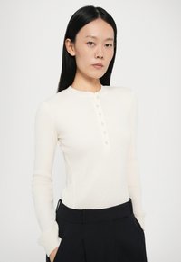 Maglione a maniche lunghe color crema con un texture a coste, caratterizzato da un colletto henley e sei bottoni, abbinato a pantaloni scuri a righe.