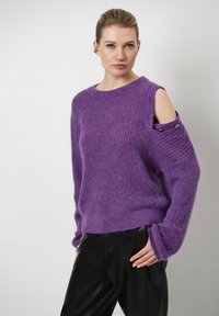 Paars gebreid vest met een losvallende pasvorm, brede mouwen en een schouderuitsnede met metallic hardware accenten. Gecombineerd met zwarte broek.