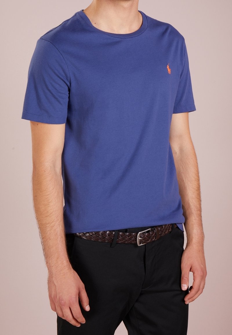 Blå bomulls-t-shirt med rund halsringning, korta ärmar och en liten broderad logotyp i orange på bröstet. Buren med svarta byxor och ett flätat bälte.