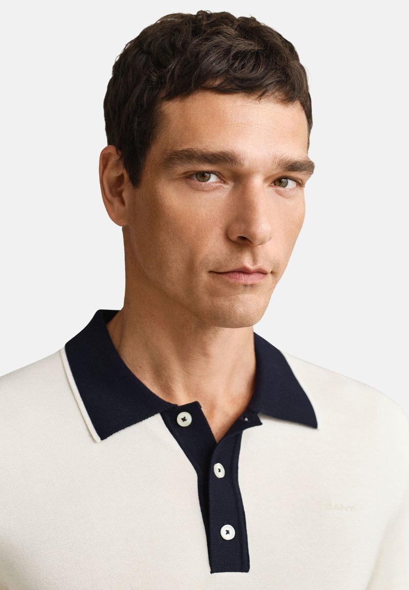 Uomo con capelli corti e scuri che indossa una polo color crema con colletto e pattina con bottoni neri, che guarda leggermente di lato.