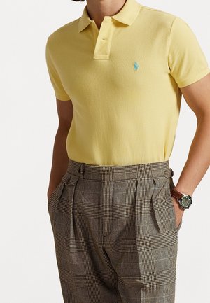 Geel poloshirt met een textuur gebreid, voorzien van een turquoise logo. Gecombineerd met bruin geruite high-waisted broek en een horloge.