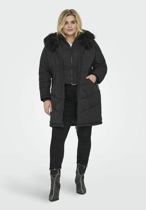 Giacca puffa nera con cappuccio in pelliccia sintetica, design trapuntato, chiusura frontale con zip e tasche laterali. Abbinata a pantaloni neri aderenti e stivali texturizzati.