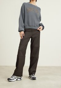 Person som bär en grå sweatshirt med glittrande text, svarta byxor och sneakers, som står mot en neutral bakgrund.