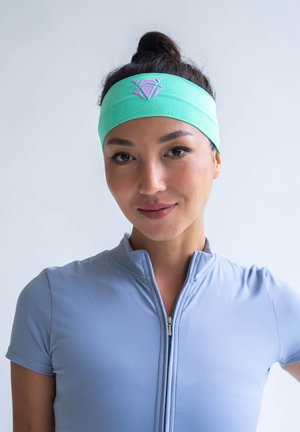 A mint green headband made of stretchy material features a purple diamond logo. The model wears a light blue zippered top.