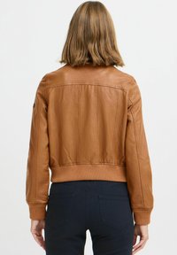 Donna con capelli castani chiari a spalla, indossa una giacca in pelle color tan corta e pantaloni scuri, vista da dietro.