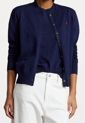 Personne portant un cardigan boutonné bleu marine par-dessus un haut assorti de la même couleur et un pantalon blanc, avec un logo rouge brodé sur la poitrine.