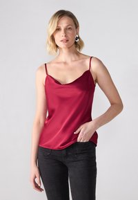 Camisola bordeaux in tessuto satinato con spalline sottili, scollatura a barchetta e vestibilità comoda, abbinata a jeans neri slim-fit.