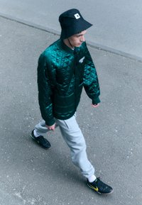 Zelená prešívaná bomber bunda, šedé tepláky, čierny klobúk typu bucket a čierne športové topánky so žltými akcentmi; chôdza po šedej ploche.