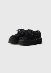 Zwarte schoenen met een donzige bovenkant en dikke zwarte rubberen plateauzool. Het ontwerp omvat vetersluiting en gouden accenten.