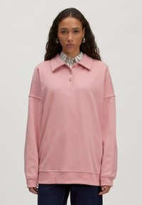 Lichtroze sweatshirt met een kraag, drie-knopen placket en verlaagde schouders. Heeft ribgebreide manchetten en zoom, gedragen over een gestreept shirt.