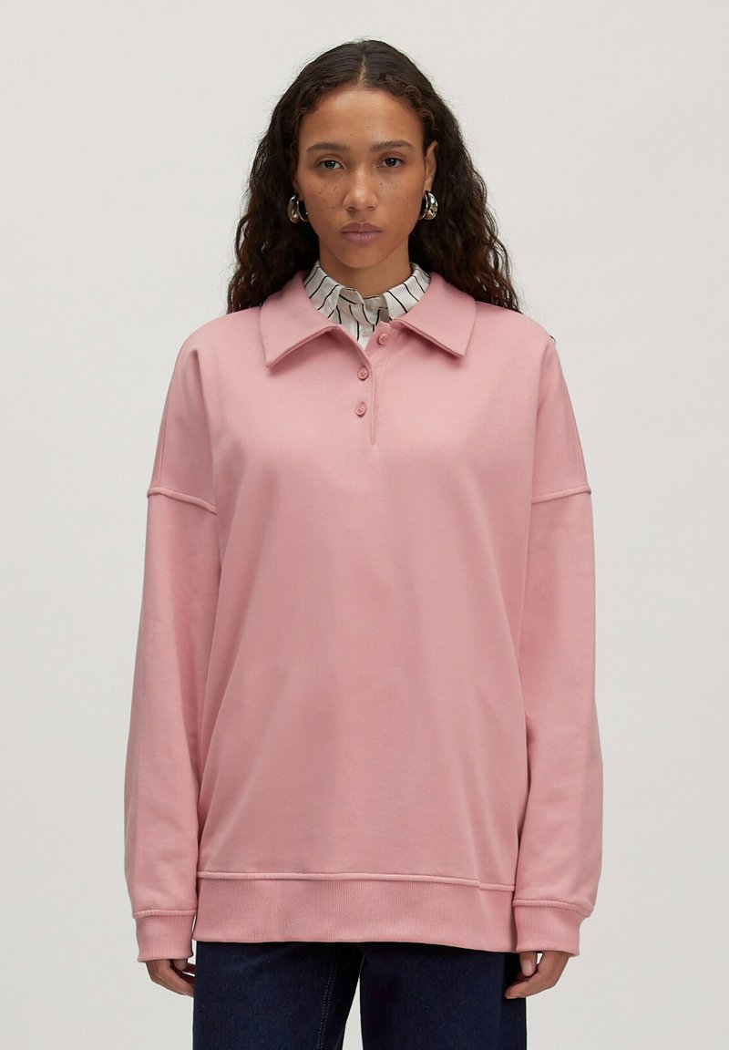 Lichtroze sweatshirt met een kraag, drie-knopen placket en verlaagde schouders. Heeft ribgebreide manchetten en zoom, gedragen over een gestreept shirt.