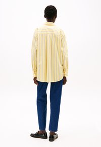 Camicia a righe gialle e bianche, vestibilità larga, maniche lunghe, yoke posteriore, abbinata a jeans blu e scarpe nere, vista da dietro.
