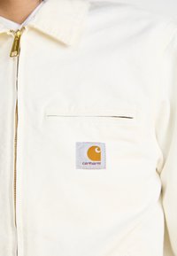 Witte katoenen jas met een rits aan de voorkant, een borstzak en een Carhartt-logo label in oranje en grijs.