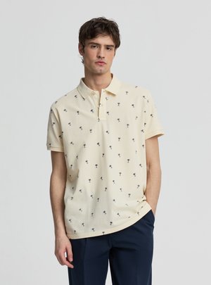 Młody mężczyzna ubrany w jasnożółtą koszulkę polo z małymi nadrukami palm i ciemnoniebieskie spodnie, stojący z jedną ręką w kieszeni.