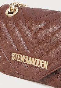 Borsa a tracolla in finta pelle marrone con texture e cuciture a zig-zag, dotata di catena dorata e accento con logo. Forma rettangolare compatta.