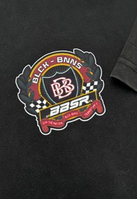 Zwarte t-shirt met een kleurrijke geborduurde patch met een schild, bloemaccenten en een geblokt patroon, waarop "BLCK - BNNS" en "BBSR" staat.