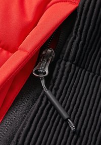Gros plan d'une fermeture éclair noire avec cordon tressé sur tissu côtelé noir à côté d'un tissu matelassé rouge vif, mettant en valeur les textures textiles et les détails de la fermeture éclair.