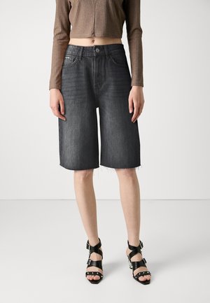 Frau trägt hochtaillierte schwarze Denim-Bermuda-Shorts mit rohem Saum, ein kurzes braunes Langarmshirt und schwarze Riemchenabsatzschuhe mit Nieten.