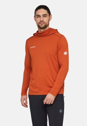 Mand med skæg og hår trukket tilbage iført en orange langærmet hættetrøje og sorte bukser mod en ensfarvet baggrund.