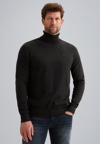 Maglione a collo alto nero realizzato in tessuto morbido, con un piccolo logo testurizzato sul lato sinistro del petto e polsini e orlo aderenti. Abbinato a jeans blu.