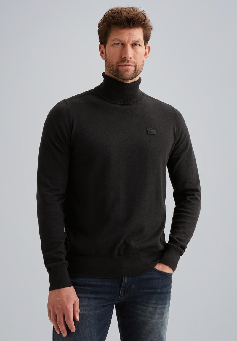 Maglione a collo alto nero realizzato in tessuto morbido, con un piccolo logo testurizzato sul lato sinistro del petto e polsini e orlo aderenti. Abbinato a jeans blu.