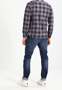 Camisa de manga larga a rayas en azul y gris, combinada con jeans de mezclilla oscuros. Zapatillas blancas con detalles en negro. Estilo casual, vista trasera visible.