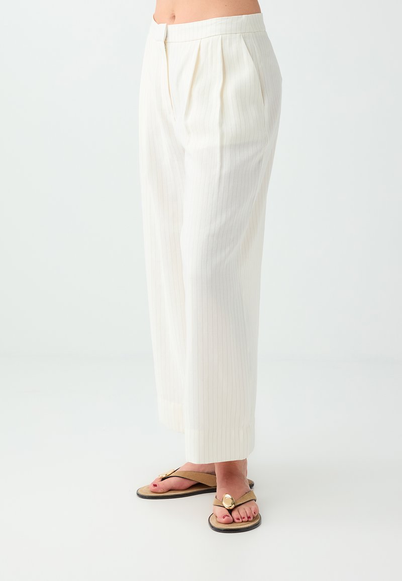 Femme portant un pantalon blanc à fines rayures, taille haute et jambes larges, avec des sandales beiges ornées de grandes décorations circulaires dorées.